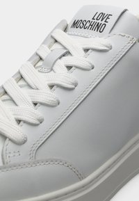 Κοντινό πλάνο λευκού αθλητικού παπουτσιού με λευκές κορδόνια και την επιγραφή "LOVE MOSCHINO" στη γλώσσα, δείχνοντας λεπτομερή ραφές στο λείο δερμάτινο πάνω μέρος.