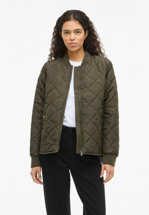 Femme aux cheveux bouclés porte une veste matelassée vert olive par-dessus une chemise blanche et un pantalon noir, debout devant un fond clair uni.