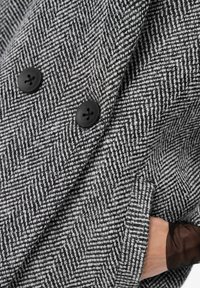 Grijs houndstooth-gepatenteerd jasje met grote zwarte knopen, voorzien van een textuurstof en een zijzak die zichtbaar is nabij de manchetten.