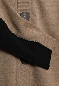 Sweter z dzianiny z beżowym korpusem o prążkowanej fakturze, czarnymi akcentami na rękawach i okrągłym czarnym guzikiem.