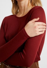 Burgundy långärmad topp med rund halsringning och texturerad tyg, figursydda ärmar och en avslappnad passform, i kombination med en rutig kjol.