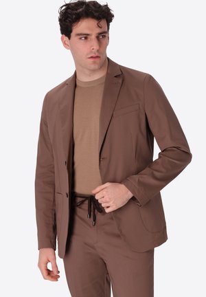 Uomo che indossa giacca e pantaloni marroni con vita con coulisse, abbinati a un maglione beige, guarda verso destra.