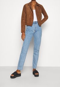 Hnědá semišová motocyklová bunda, světlý tílko, světlé rovné džíny modré barvy a černé platformové sandály s žebrovanými popruhy.