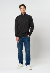 Črni pulover s polovičnim zadrgo, narejen iz pletenine, v kombinaciji z modrimi jeans hlačami in belimi supergami. Ima majhen emblem na prsih.