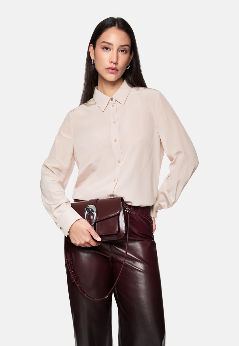 Beige long-sleeve shirt with button front, worn with glossy burgundy faux leather pants and a small matching handbag featuring a silver clasp.