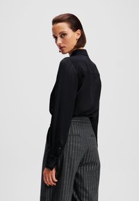 Camicia in satin nera con colletto e polsini abbottonati, abbinata a pantaloni grigi a righe sottili con vita aderente e tasche.