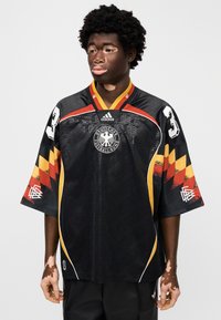 Μαύρη φανέλα με κοντά μανίκια και κόκκινες, κίτρινες και πορτοκαλί λεπτομέρειες, με γεωμετρικό μοτίβο, στρογγυλή λαιμόκοψη και το λογότυπο της Γερμανικής Ποδοσφαιρικής Ομοσπονδίας.