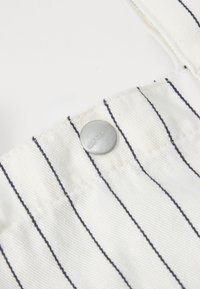 Tela blanca con rayas verticales en azul marino, que presenta un botón de metal plateado con la inscripción "carhartt" grabada. Primer plano de la textura de la ropa y detalles de diseño.