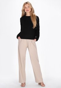 Zwarte gebreide trui met ronde hals en lange mouwen, gecombineerd met beige broek met wijde pijpen en lichte sandalen. Soepele texturen en strakke lijnen.