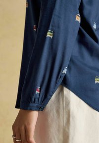 Navy blue long-sleeve shirt with small colorful car patterns, worn with light beige pants, hand with silver rings visible.