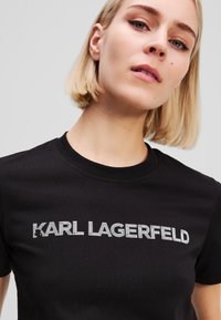 T-shirt nera in cotone con testo in glitter argento "KARL LAGERFELD" sul petto. Collo rotondo, maniche corte, texture liscia.