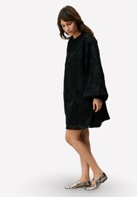 Robe noire à texture avec une coupe ample, des manches longues et un col rond. Associée à des chaussures plates argentées métallisées avec un détail de boucle.