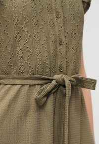 Κοντινό πλάνο υφασμάτινου φόρεματος σε ελιά πράσινο χρώμα με κουμπιά μπροστά και δέσιμο με υφασμάτινη ζώνη στη μέση.