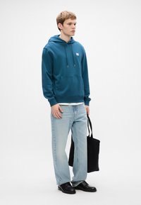 Žalsvai mėlyna, su gobtuvu, palaidinė su priekinėmis kišenėmis, derinama su šviesiai mėlynais tiesiais džinsais ir juodais batais. Juoda rankinė laikoma dešinėje rankoje.