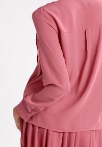 Long-sleeve blouse in soft pink fabric, smooth texture, slightly loose fit, featuring a back pleat and button detail on the sleeve cuff.