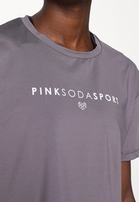 Närbild på en person som bär en grå T-shirt med texten "PINK SODA SPORT" och en geometrisk hjärtlogotyp tryckt i vitt på bröstet.