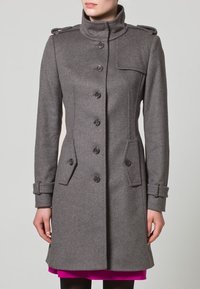 Harmaa villakangastakki, jossa korkea kaulus, napitettava suljenta, sivutaskut ja olkatoppaukset; takissa on istuva leikkaus ja teksturoitu kangas.