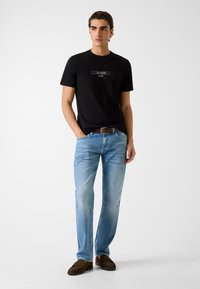 Zwart katoenen T-shirt met geperst "GUESS"-logo, lichtblauwe spijkerbroek en bruine suède mocassinschoenen. Eenvoudige, casual outfit.