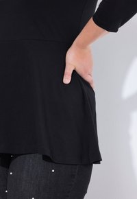 Schwarzes Tunika-Oberteil aus weichem Stoff mit abgerundetem Saum und seitlichen Taschen. Verfügt über Dreiviertelärmel und wird mit dunkler Jeans getragen, die mit Perlen verziert ist.