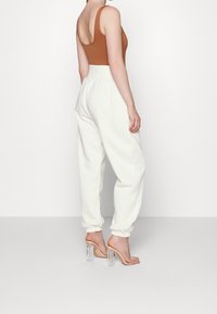 Žena oblečená v vysokých bílých joggerech, bezramínkovém hnědém topu a průhledných vysokých podpatcích, stojící na jednobarevném bílém pozadí.