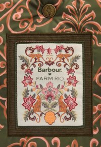 Zielony materiał z różowymi kwiatowymi wzorami ma prostokątną metkę. Metka wyświetla napisy "Barbour" oraz "FARM RIO" z kolorowym haftem przedstawiającym kwiaty i zwierzęta.