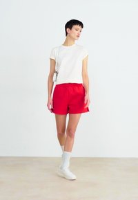 Balta trumpa rankovė t-shirt su apvalia iškirpte, derinama su ryškiomis raudonomis šortais ir baltais sportbačiais. Modelis taip pat neša mažą per petį nešiojamą maišelį.