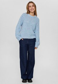 Hellblauer, gestrickter Pullover mit Rippenkragen und langen Ärmeln, der bunte Farbspritzer aufweist, kombiniert mit weiten, dunklen Jeanshosen.