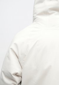 Isik kannab heledat kapuutsiga jakk, mille tagumise ja õlavarre detailid on nähtavad lihtsa valge tausta taustal.