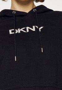 Personne portant un sweat à capuche noir avec le logo DKNY blanc centré sur la poitrine et des cordons noirs avec embouts métalliques.