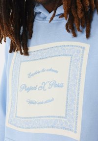 Felpa blu chiaro con cappuccio, caratterizzata da un grande grafico rettangolare bianco con intricati bordi paisley blu chiaro e la scritta "Project X Paris."