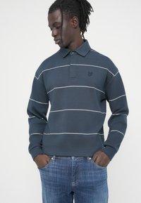 Μακρυμάνικο πόλο σε ναυτικό μπλε χρώμα με λευκές οριζόντιες ρίγες, που διαθέτει γιακά και μικρό κεντητό λογότυπο στο στήθος. Φορεμένο με τζιν.