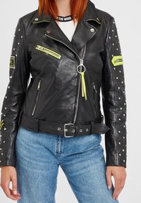 Černá kožená moto bunda s kovovými cvočky, neonovými akcenty a několika zipy. Pas s opaskem a texturovaný povrch zvýrazňují její design.