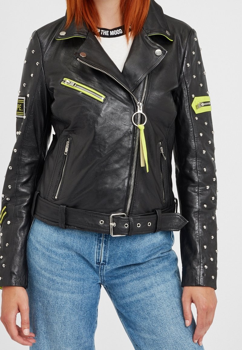 Černá kožená moto bunda s kovovými cvočky, neonovými akcenty a několika zipy. Pas s opaskem a texturovaný povrch zvýrazňují její design.