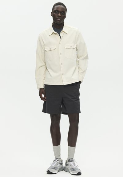 Jeune homme portant une veste crème boutonnée, un short noir, des chaussettes grises et des baskets grises, debout devant un fond blanc uni.