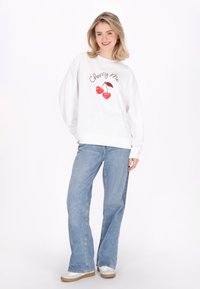 Felpa bianca con testo "Cherry Me" e grafica di ciliegia rossa, abbinata a jeans a gamba larga blu chiaro e sneakers bianche.