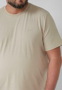 Beige Baumwoll-T-Shirt mit rundem Halsausschnitt und kurzen Ärmeln. Mit dezentem Branding in passendem Faden. Glatte Textur.