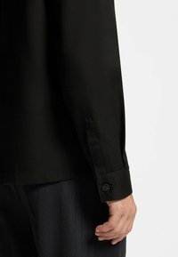 Chemise noire à manches longues avec un poignet boutonné, présentant une texture lisse et une coupe ajustée, associée à un pantalon sombre à fines rayures.
