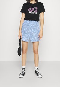 Svart t-shirt med rosa grafisk design och texten "Converse", tillsammans med ljusblå shorts med ett vitt blommönster, bärs med svarta hög topp sneakers.