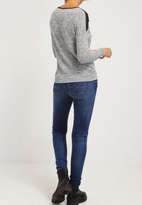 Haut long gris texturé avec des accents en dentelle noire sur les épaules, associé à un jean bleu ajusté et des bottines noires.