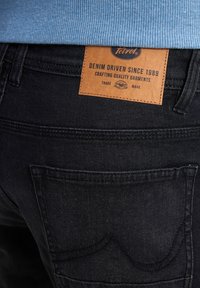 Zwarte denim jeans hebben een gestructureerd leren label met de tekst "Petrol," dat vermeldt "Denim Driven Since 1989." Inclusief stiksels op de achterzak.