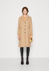 Beige ullfrakk med spiss krage, fem mørke knapper og to frontlommer. Kombinert med svarte leggings og en gullveske.