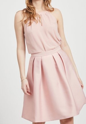 Vrouw in een mouwloze lichtroze geplooide jurk met een sleutelgat-halslijn en een aangesloten tailleband, staand tegen een effen achtergrond.