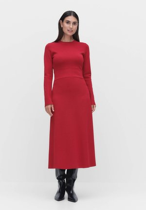 Röd stickad klänning med långa ärmar, figurnära bodice och flödande kjol. Bärs med glansiga svarta knähöga stövlar. Slät textur, minimalistisk design.
