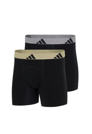 Athletic man standing barefoot, shirtless, wearing black boxer briefs with a beige waistband featuring a black logo.