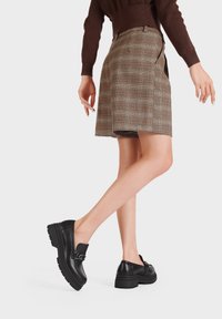 Scarpe platform in pelle nera con suola spessa e una fibbia decorativa. Indossate con una gonna scozzese marrone, evidenziando un design elegante.