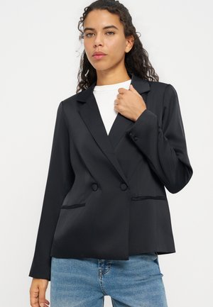 Jeune femme aux cheveux bouclés porte un blazer noir croisé sur un haut blanc et un jean bleu, tenant le revers du blazer d'une main.
