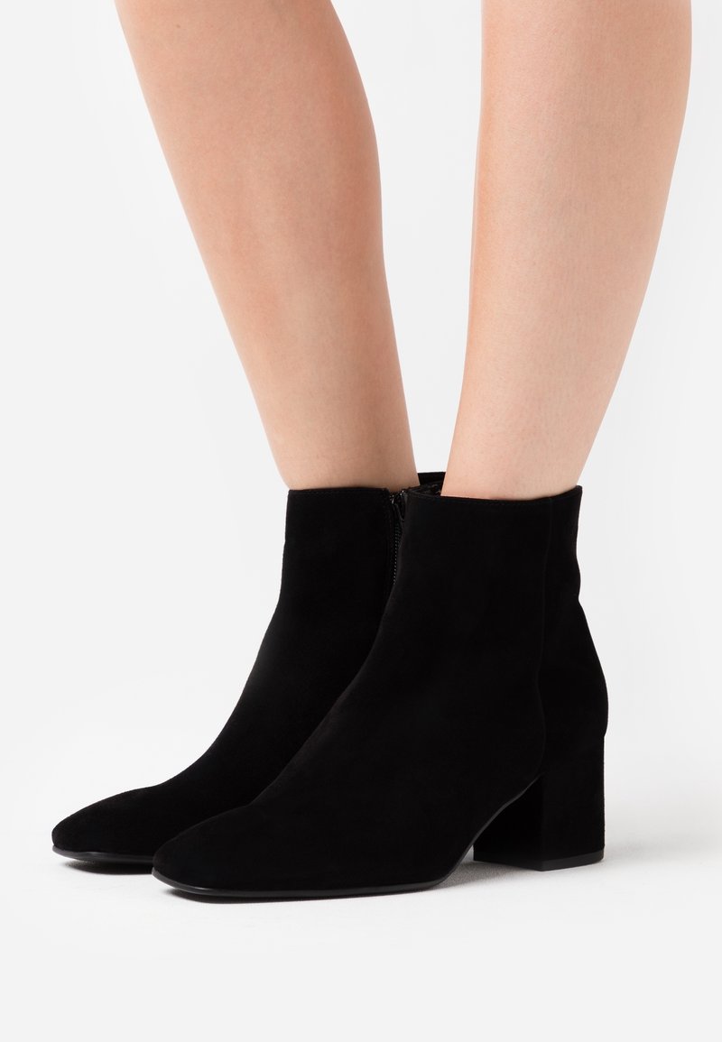 Bottines en suède noir à talons carrés portées sur des jambes nues sur fond blanc.