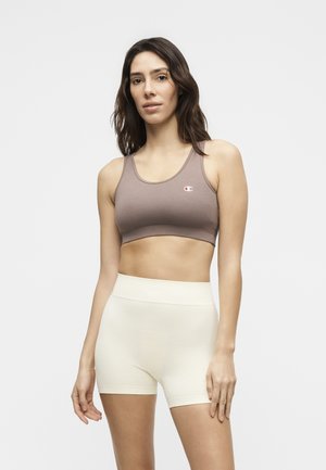Kobieta w mauve sportowym biustonoszu i wysokich kremowych, teksturowanych krótkich spodenkach rowerowych, stojąca na jasnym, jednolitym tle.