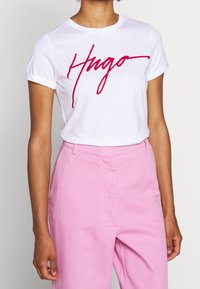 Hvid bomulds T-shirt med rød kursiv "Hugo" logo, korte opsmøgede ærmer, parret med lyserøde højtaljede bukser. Glat tekstur.