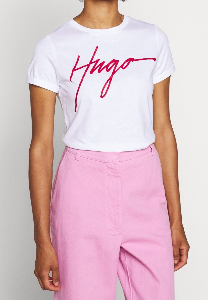 Hvid bomulds T-shirt med rød kursiv "Hugo" logo, korte opsmøgede ærmer, parret med lyserøde højtaljede bukser. Glat tekstur.